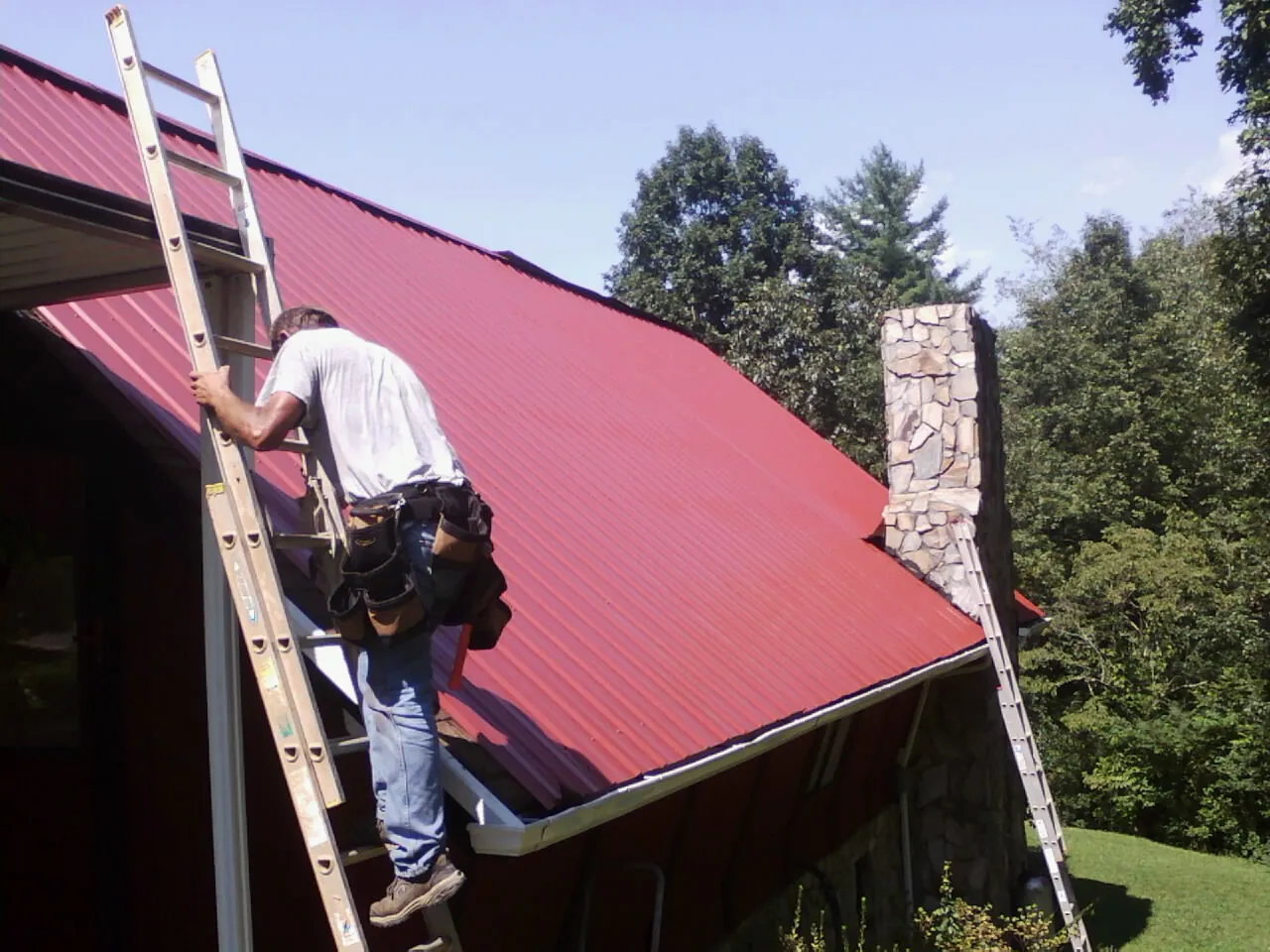 Roofer on a ladder beside a red metal roof and stone chimney — Soffit & Fascia Repair by Roofing Brothers in Fairmont